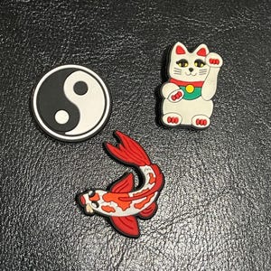 Może przedstawiać: Trzy ozdobne zawieszki: czarno-biały symbol Yin Yang, biały szczęśliwy kot z czerwoną obrożą oraz czerwono-biały karp koi. Te zawieszki są prawdopodobnie przeznaczone do butów lub akcesoriów.
