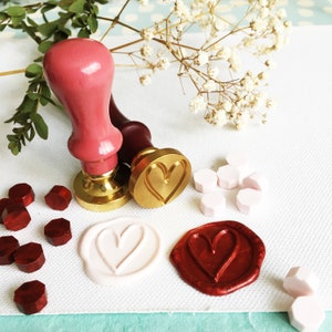 May include: Two wooden wax seal stamps with heart designs. One stamp is gold with a heart design, the other is red with a heart design. There are also wax seals in red, white, and gold.