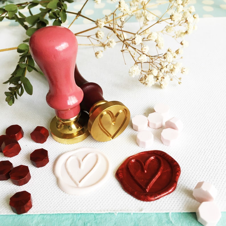 May include: Two wooden wax seal stamps with heart designs. One stamp is gold with a heart design, the other is red with a heart design. There are also wax seals in red, white, and gold.