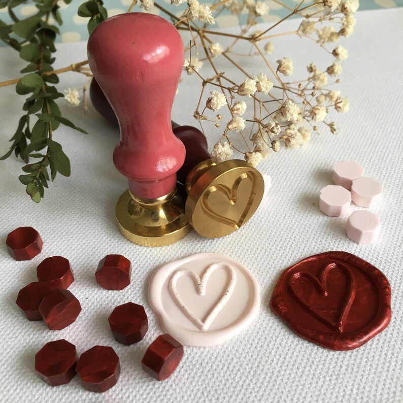 May include: A set of wax seals with a heart design. The seals are made of red, white, and gold wax. There are also several red wax seal pellets and a wooden handle with a gold base.