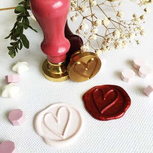 May include: A set of three wax seals with heart designs. The seals are gold, red, and pink. The seals are on a white surface with white flowers and small white and pink decorative items.