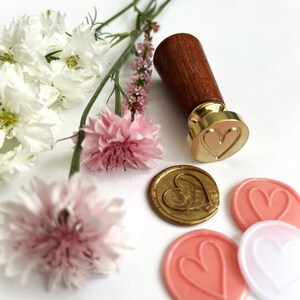 May include: A wooden handle wax seal stamp with a gold heart design. The stamp is surrounded by pink and white flowers and wax seals with heart designs.