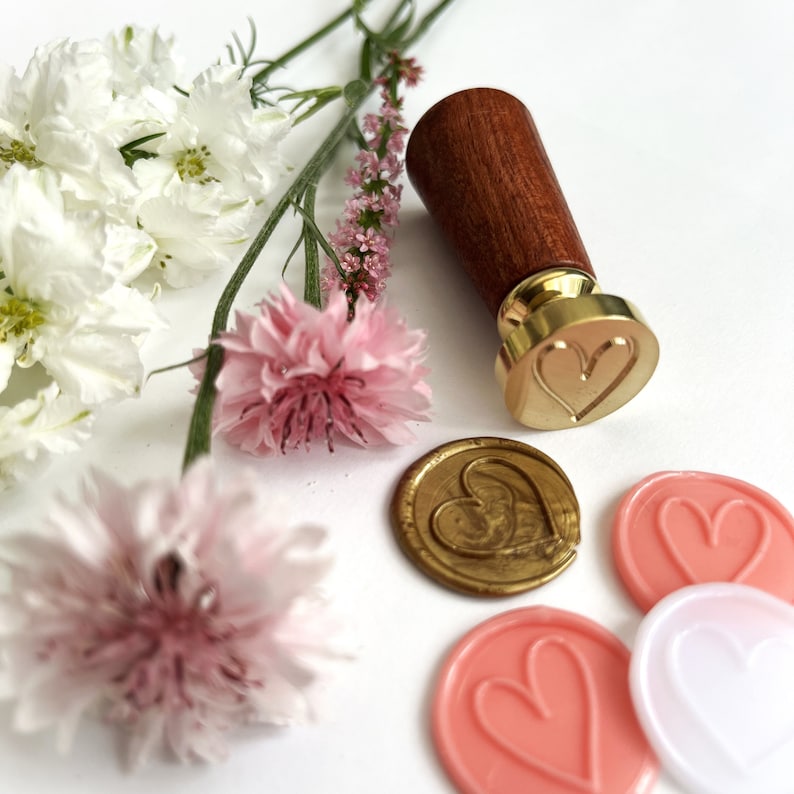 May include: A wooden handle wax seal stamp with a gold heart design. The stamp is surrounded by pink and white flowers and wax seals with heart designs.