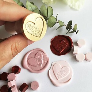 May include: A gold heart-shaped wax seal stamp with a leafy design. The stamp is being used to create wax seals in pink and red.