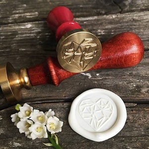 May include: A gold-toned wax seal stamp with a geometric heart design. The stamp is attached to a wooden handle with a red finish. A white wax seal with the same design is also shown.