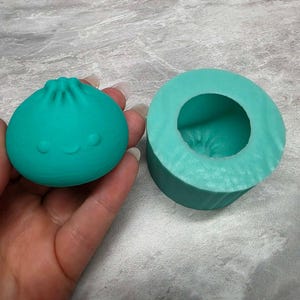 May include: A teal silicone mold and a matching teal dumpling-shaped figure with a small smiling face. The figure features a pleated top and a smooth, rounded body. The mold is cylindrical with a hollow center that mirrors the shape of the figure.