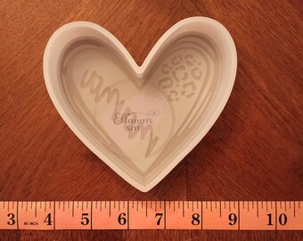 May include: White ceramic heart-shaped dish with a raised design of a swirl and leopard print. The dish is approximately 6 inches wide.