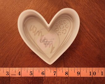 May include: White heart-shaped cookie cutter with a swirl design and a leopard print design inside. The cutter is approximately 6 inches wide.