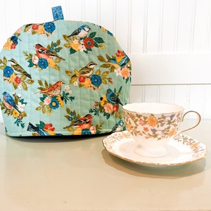 May include: A teal blue tea cozy with a quilted design and a pattern of colorful birds and flowers. The tea cozy is sitting on a white table next to a white teacup and saucer with a floral pattern.