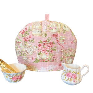 May include: A pink quilted teapot cozy with a floral pattern featuring roses, teapots, and cups. The cozy has a loop for hanging. The text "Roses" and "Flowers" is visible on the cozy.