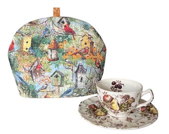 May include: A tea set featuring a birdhouse-themed tea cozy and a teacup with saucer. The cozy has a colorful bird and birdhouse pattern. The teacup and saucer have a fruit design.