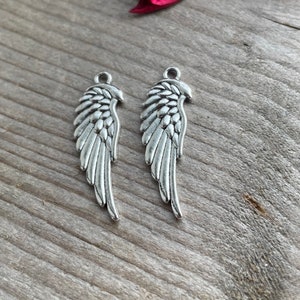 May include: Two silver angel wing charms with a detailed design. The wings are slightly curved and have a textured surface.