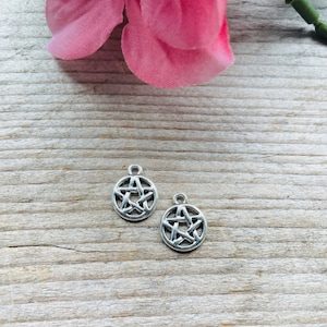 Puede incluir: Dos colgantes de pentagrama plateados con un marco circular, sobre una superficie de madera envejecida. Los colgantes presentan un diseño de estrella de cinco puntas. Una flor rosa y un tallo verde están en el fondo.