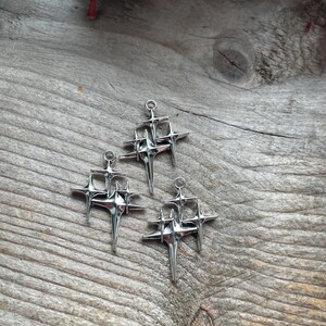 May include: Three silver-toned metal charms, each shaped like a cluster of four-pointed stars with elongated rays. Each charm features a small circular loop at the top for attachment, with a polished, reflective surface and sharp, geometric points.