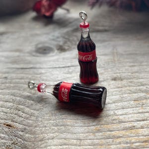 May include: Miniature Coca-Cola bottle charms with red and white labels. One bottle stands upright, the other lies on its side. Both have silver-coloured loops at the top for hanging. The bottles are on a weathered wooden surface.