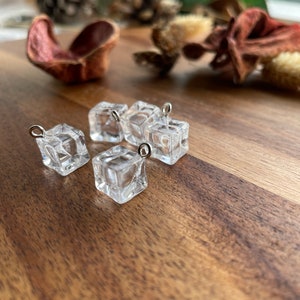 Op de afbeelding: Vier transparante plastic ijsblokjes bedels met zilveren springringen. De bedels liggen op een houten oppervlak.