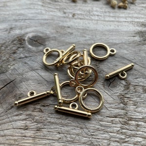 May include: A close-up of a pile of gold-toned metal toggle clasps. The clasps are in various positions, showing the different parts of the clasp, including the bar, the loop, and the ring.