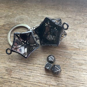 Puede incluir: Un llavero contenedor de metal oscuro con forma de estrella y bisagras que se abre para revelar varios dados poliédricos pequeños de múltiples caras en su interior. Tres dados metálicos oscuros adicionales están fuera del contenedor.
