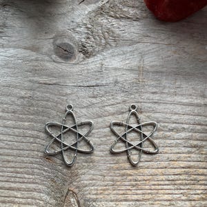 May include: Two silver atom-shaped pendants. Each pendant has a six-pointed star design with intersecting lines, and a small loop at the top for hanging. The pendants are displayed on a weathered wooden surface.
