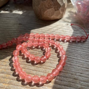 May include: Strand of faceted, light pink beads on a red string. The beads are oval-shaped and have a sparkling, translucent quality. The beads are displayed on a weathered wooden surface, with a wooden object in the background.