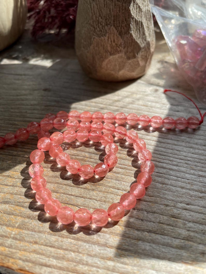 May include: Strand of faceted, light pink beads on a red string. The beads are oval-shaped and have a sparkling, translucent quality. The beads are displayed on a weathered wooden surface, with a wooden object in the background.