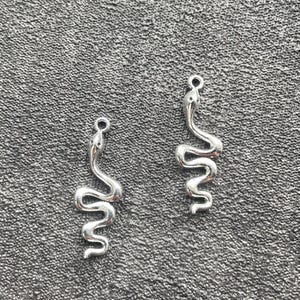 May include: Two silver-toned metal snake charms, each featuring a smooth, polished finish and a small circular loop at the top for attachment. The snake figures are depicted in a winding, S-shaped posture with a defined head and a curved tail.