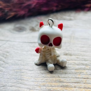 May include: A small pendant shaped like a seated skeleton figure with a white skull, red eye sockets, and red horn-like accents on top. The figure has a skeletal body and a silver-toned metal jump ring attached to the top of the head for hanging.