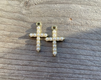 May include: Two gold-colored cross pendants, each adorned with small, round, off-white pearls. The crosses are displayed on a weathered, gray wooden surface, with the sunlight casting shadows.