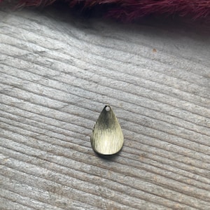 May include: A teardrop-shaped metal pendant with a brushed gold finish and a small circular hole at the top for attachment. The surface of the charm features fine vertical lines, creating a textured appearance across the metallic material.