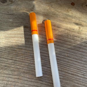 May include: Two white and orange cigarette-shaped charms with a red stripe. The charms are made of polymer clay and have a shiny finish.