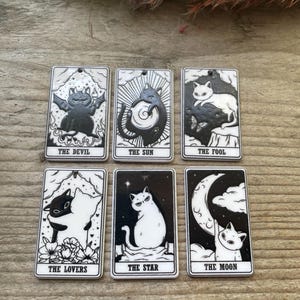 May include: Six rectangular black and white charms featuring cat-themed tarot card designs. The cards are labelled The Devil, The Sun, The Fool, The Lovers, The Star, and The Moon. Each charm has a small hole at the top for hanging as a pendant.