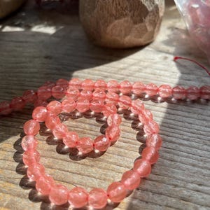 May include: A strand of faceted, light pink gemstone beads, likely rose quartz, arranged in a spiral on a wooden surface. The beads are round and have a subtle sparkle. A wooden carved object is in the background.