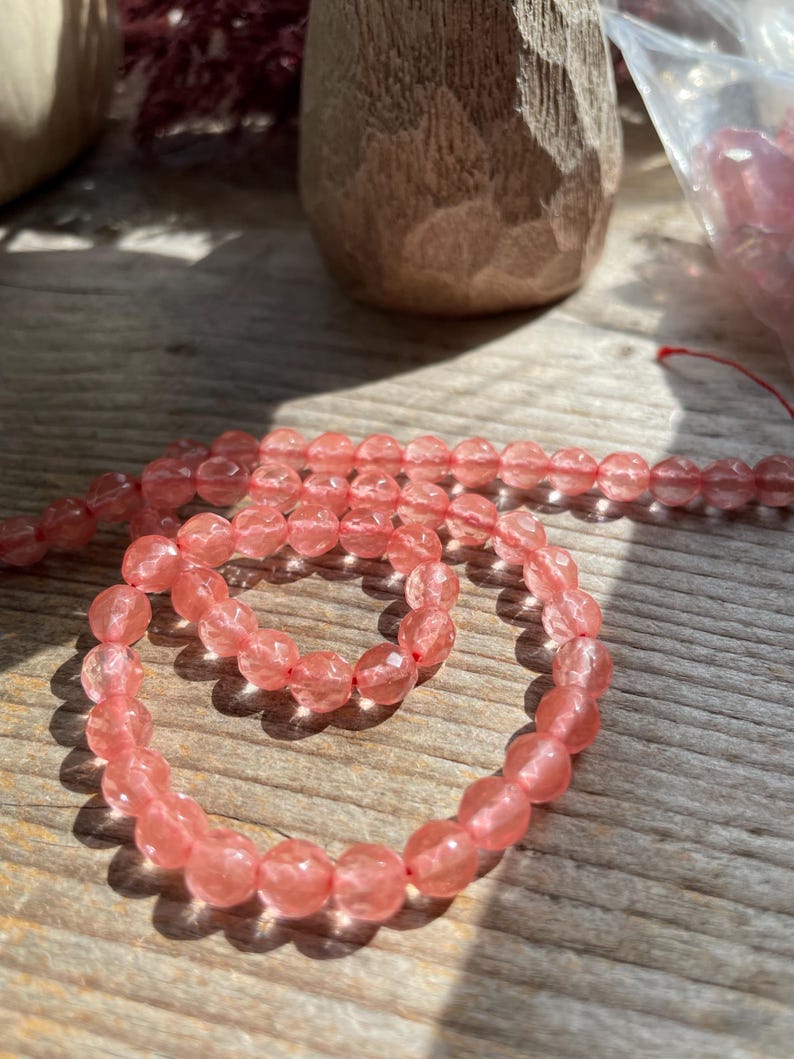 May include: A strand of faceted, light pink gemstone beads, likely rose quartz, arranged in a spiral on a wooden surface. The beads are round and have a subtle sparkle. A wooden carved object is in the background.