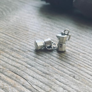 Puede incluir: Dos dijes de metal plateado con forma de cafeteras espresso en miniatura. Un dije está colocado horizontalmente mientras que el otro está de pie, ambos con detalles de asas, tapas y boquillas en un acabado metálico pulido.