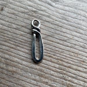 May include: A silver-toned metal pendant featuring an elongated, teardrop-shaped loop with a twisted, knot-like design at the top. The pendant has a smooth, polished finish and a small circular opening at the very top for attachment to a chain or cord.