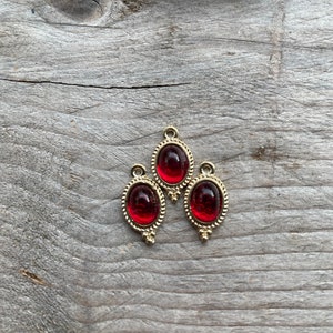Op de afbeelding: Drie goudkleurige ovale bedels met rode glazen cabochons. De bedels hebben een decoratieve rand en zijn perfect voor het maken van sieraden.