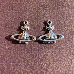 May include: Two silver-colored planet-shaped charms with a cross on top, set against a patterned purple background. The charms have a ring at the top for attaching to a necklace or bracelet. The design is detailed and symmetrical.