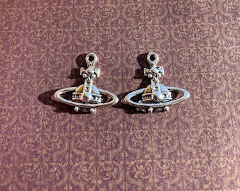 May include: Two silver-colored planet-shaped charms with a cross on top, set against a patterned purple background. The charms have a ring at the top for attaching to a necklace or bracelet. The design is detailed and symmetrical.
