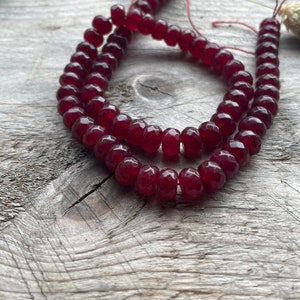 May include: A strand of deep red, faceted, circular beads arranged in a loose coil. The beads have a translucent quality and a uniform, polished finish, strung together on a thin, dark red thread that is visible between the individual pieces.