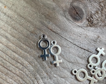 Puede incluir: Primer plano de varios abalorios de metal plateado en forma de símbolo femenino. Los abalorios están esparcidos sobre una superficie de madera.