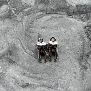 Op de afbeelding: Twee zilveren tandcharms met een lus aan de bovenkant om aan sieraden of andere knutselwerken te bevestigen.