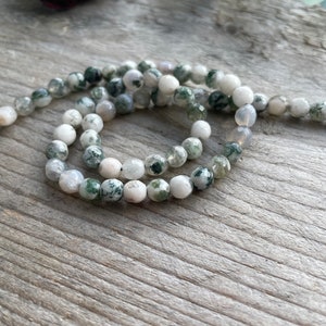 Könnte beinhalten: Stränge aus runden, polierten Edelsteinperlen in Weiß- und Grüntönen. Die Perlen haben ein gesprenkeltes Muster und sind zu einer Halskette oder einem Armband aufgereiht. Die Perlen werden auf einer Holzoberfläche präsentiert.