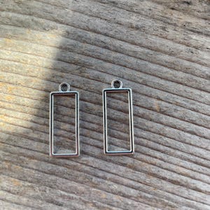 May include: Two silver-colored rectangular pendant frames with a small loop at the top for attaching to a chain or cord. The frames are empty and sit on a weathered wooden surface.