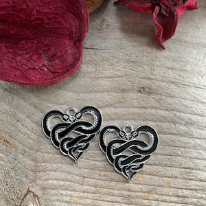 May include: Two silver heart-shaped charms with black enamel snake designs. The snakes are intertwined and form a heart shape.