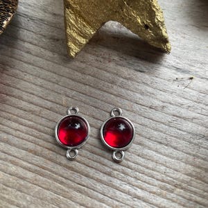 Pode incluir: Dois pingentes de metal prateado com cabochões de vidro vermelho. Cada pingente tem um laço de cada lado para prender em joias.