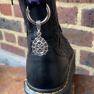 May include: Black leather boot with a silver spiderweb charm. The charm is a teardrop shape with a spider and web design. The boot has a purple strap with white text and yellow stitching.