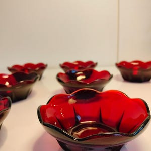 Könnte beinhalten: Eine Sammlung rubinroter Glasschalen in Blütenform. Jede Schale hat einen dunklen Rand und einen facettierten Boden. Die Schalen sind auf einer weißen Oberfläche angeordnet, um ihre leuchtende Farbe und ihr Design hervorzuheben.