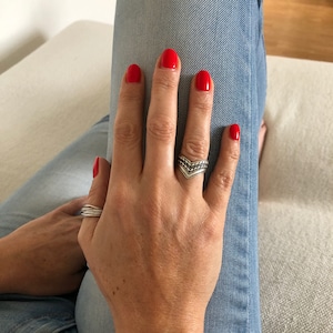 Peut inclure: Une main avec du vernis à ongles rouge porte une bague en argent avec un motif en chevron. La main repose sur un jean bleu.