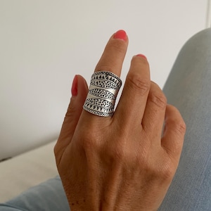 Könnte beinhalten: Ein breiter, silberfarbener Ring mit einem komplizierten Muster aus Dreiecken, Punkten und geschwungenen Linien. Der Ring wird an einem Finger getragen und zeigt sein detailliertes Design. Die Oberfläche des Rings reflektiert das Licht und hebt seine Textur und Verarbeitung hervor.