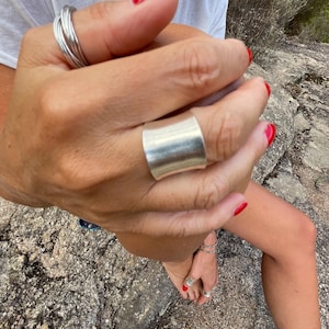 May include: Several silver rings are displayed on the fingers and toes. A broad, curved silver ring is the main feature, alongside multiple thin bands on the fingers. The toenails are painted red, and the skin tone is fair.